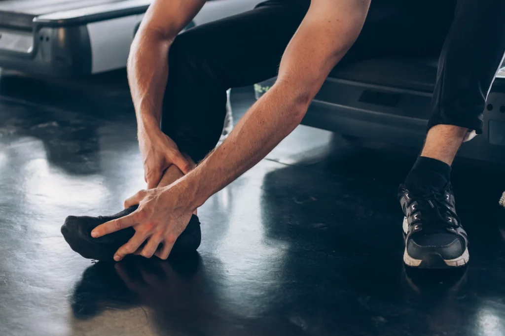 Male athlete sitting on gym floor clutching injured ankle with both hands